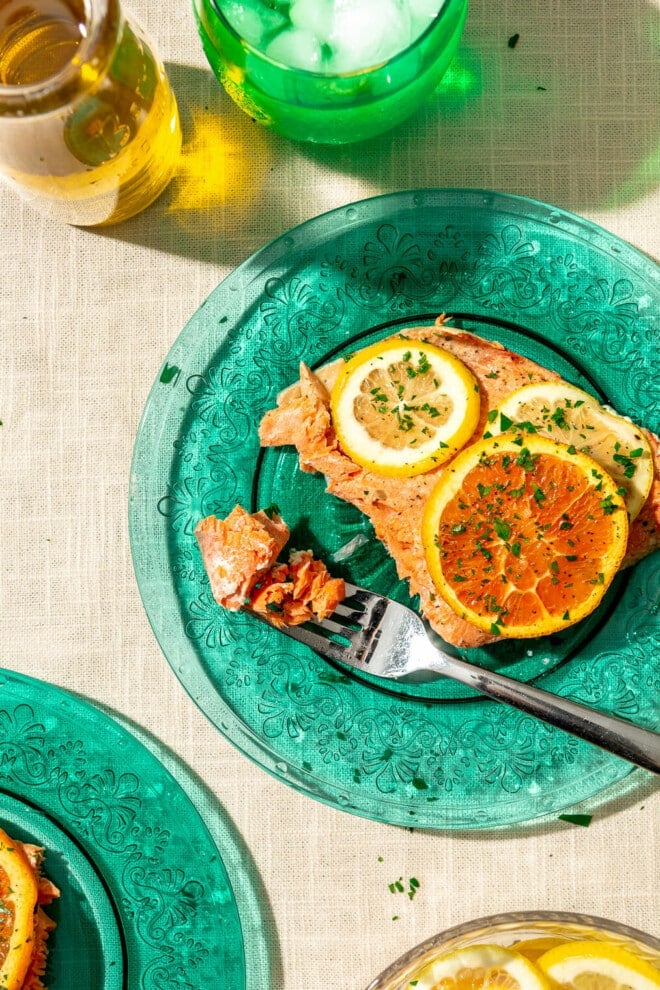 Sliced salmon filet topped with orange and lemon slices and a sprinkle of chopped parsley on translucent teal plates on a linen covered surface. There is a green glass with ice water and a glass bowl with sliced citrus on the table.