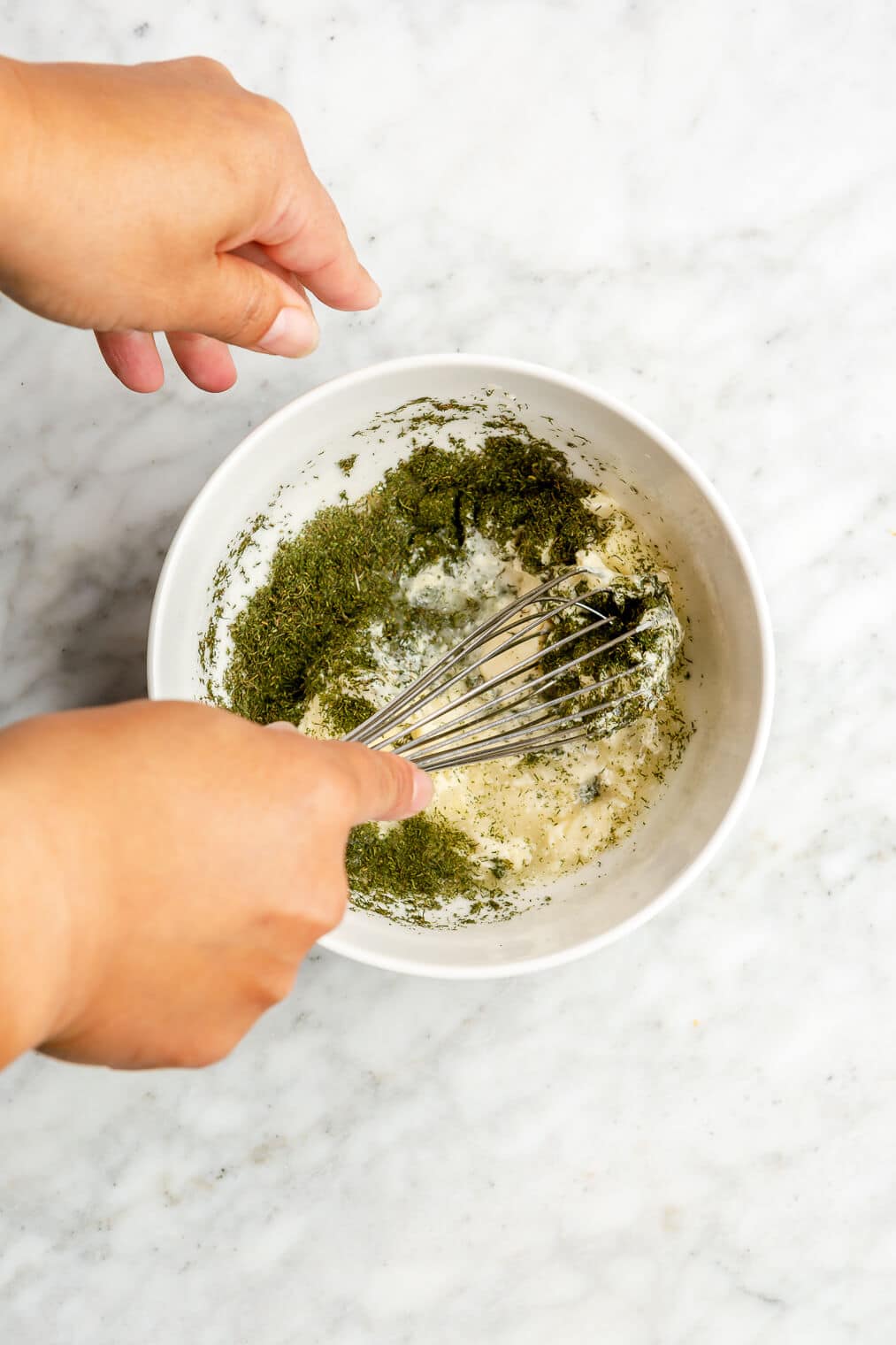 Hand whisking together mayonnaise, lemon juice, and dill in a small white bowl on a grey and white marble surface.