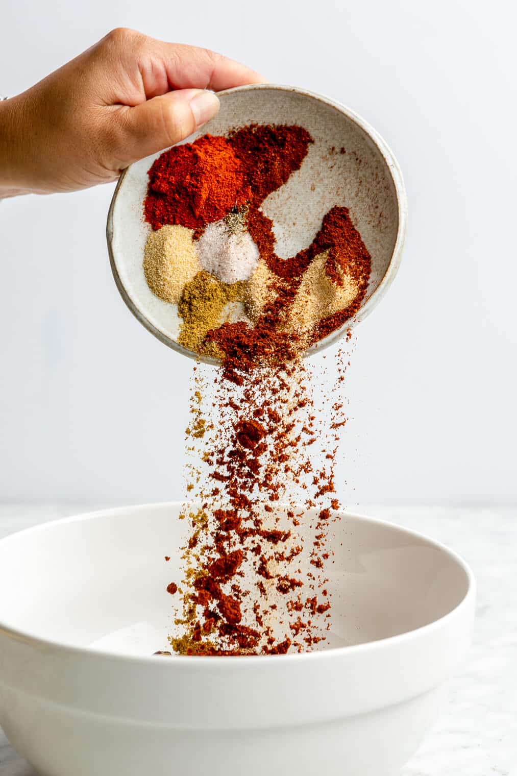 A hand pouring a small plate full of spices into a white bowl sitting on a grey and white marble surface with a white background.