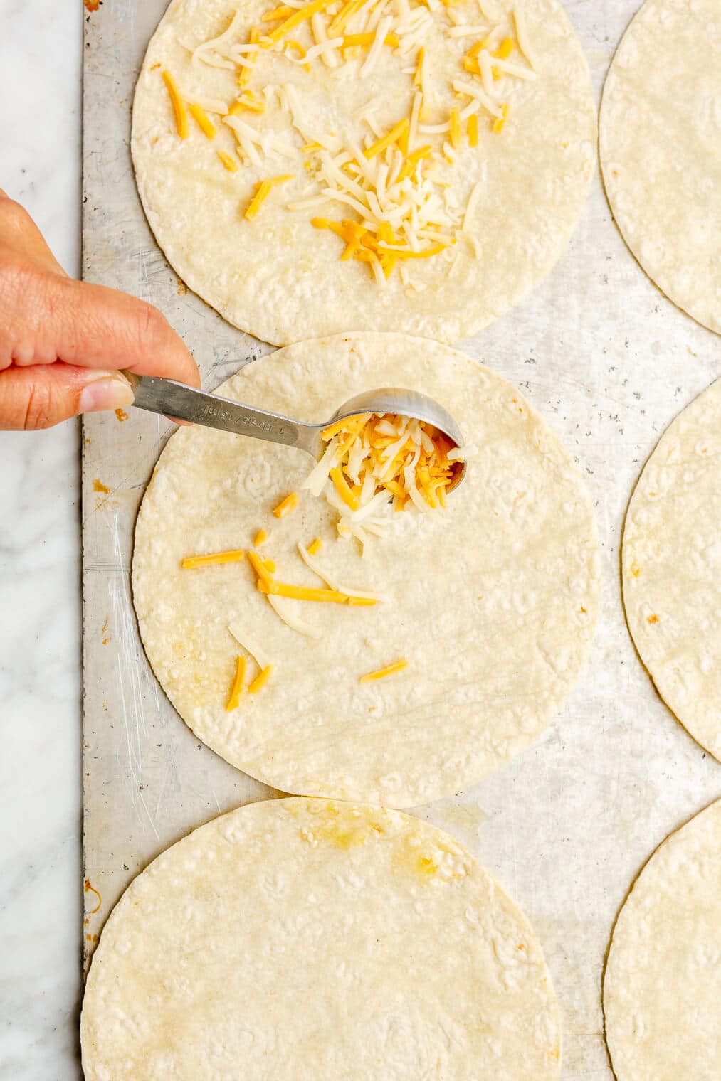 Hand scooping a tablespoon of shredded cheese onto a corn tortilla. The corn tortilla is on a metal sheet pan with 5 other corn tortillas.