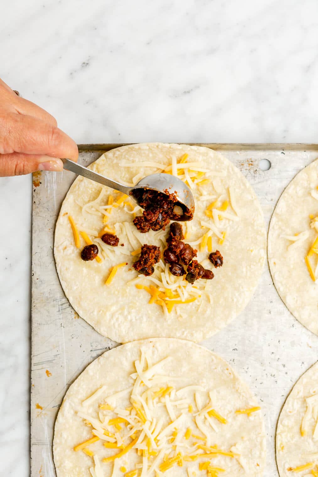 A hand scooping a tablespoon of black beans over top of shredded cheese on a corn tortilla on a metal sheet pan.