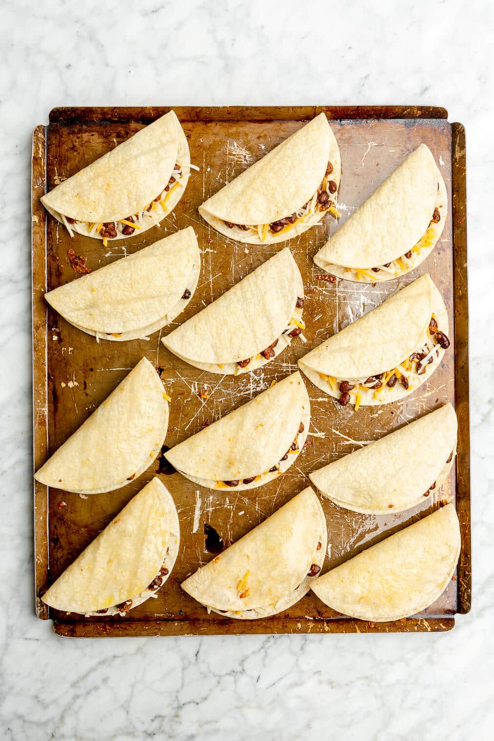 Top down of a metal sheet pan with 12 tortillas filled with beans and cheese folded in half.