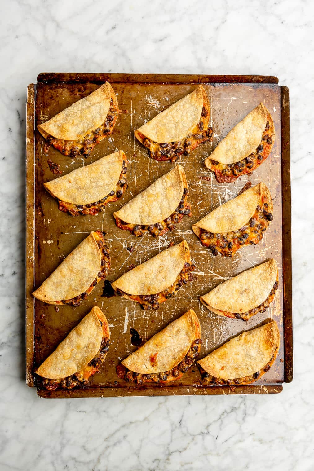 Top down of a metal sheet pan with 12 tortillas filled with beans and cheese folded in half, crisped, and baked.