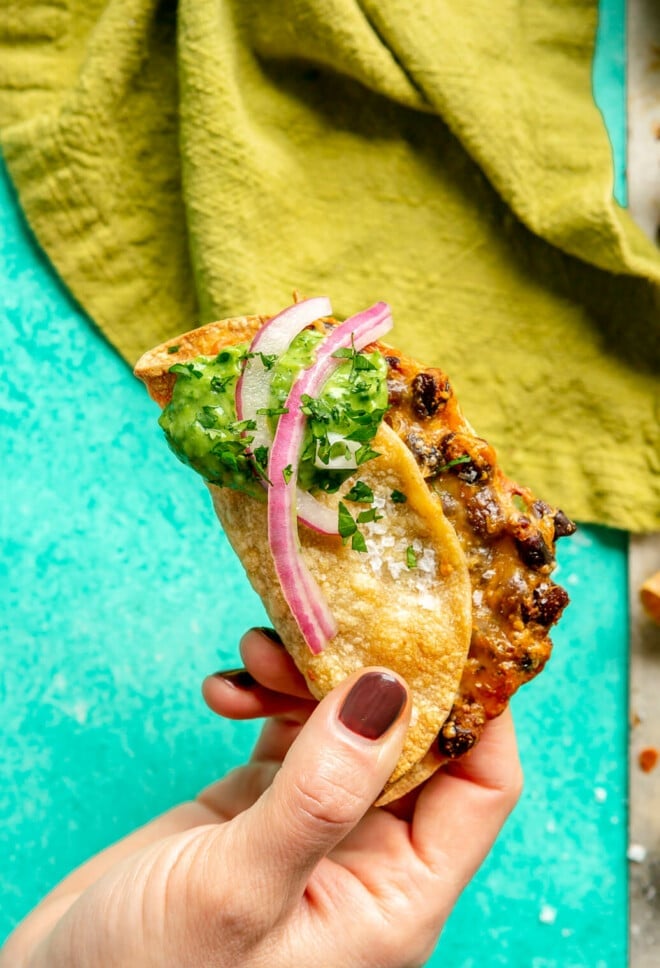 A hand holding a black bean and cheese taco topped with a dollop of a creamy, green sauce and a couple pink pickled onions held over a teal surface with a green linen draped in the background.
