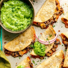 A metal sheet pan with crispy bean and cheese tacos. There is a metallic bowl with a bright green, creamy, avocado sauce and a wooden bowl with pink pickled onions. There is a green linen draped to the side. Some of the tacos are topped with a dollop of green sauce and a couple slices of pink pickled onions.