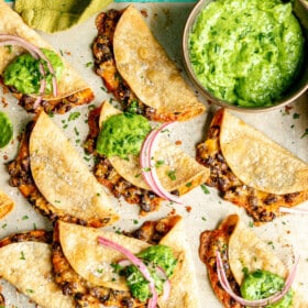 A metal sheet pan with crispy bean and cheese tacos. There is a metallic bowl with a bright green, creamy, avocado sauce and a wooden bowl with pink pickled onions. There is a green linen draped to the side. Some of the tacos are topped with a dollop of green sauce and a couple slices of pink pickled onions. All are sitting on a teal surface.