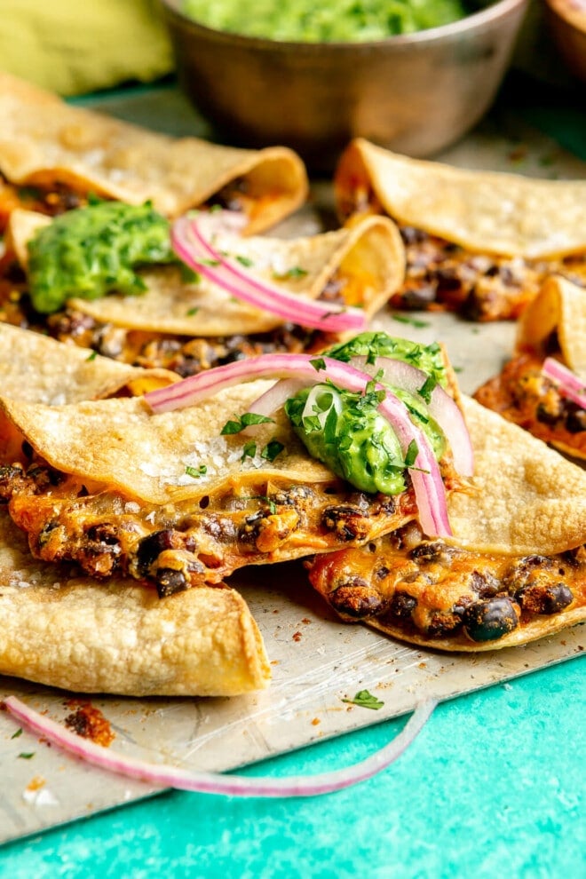 A metal sheet pan with crispy bean and cheese tacos. There is a metallic bowl with a bright green, creamy, avocado sauce. Some of the tacos are topped with a dollop of green sauce and a couple slices of pink pickled onions and flaky sea salt.