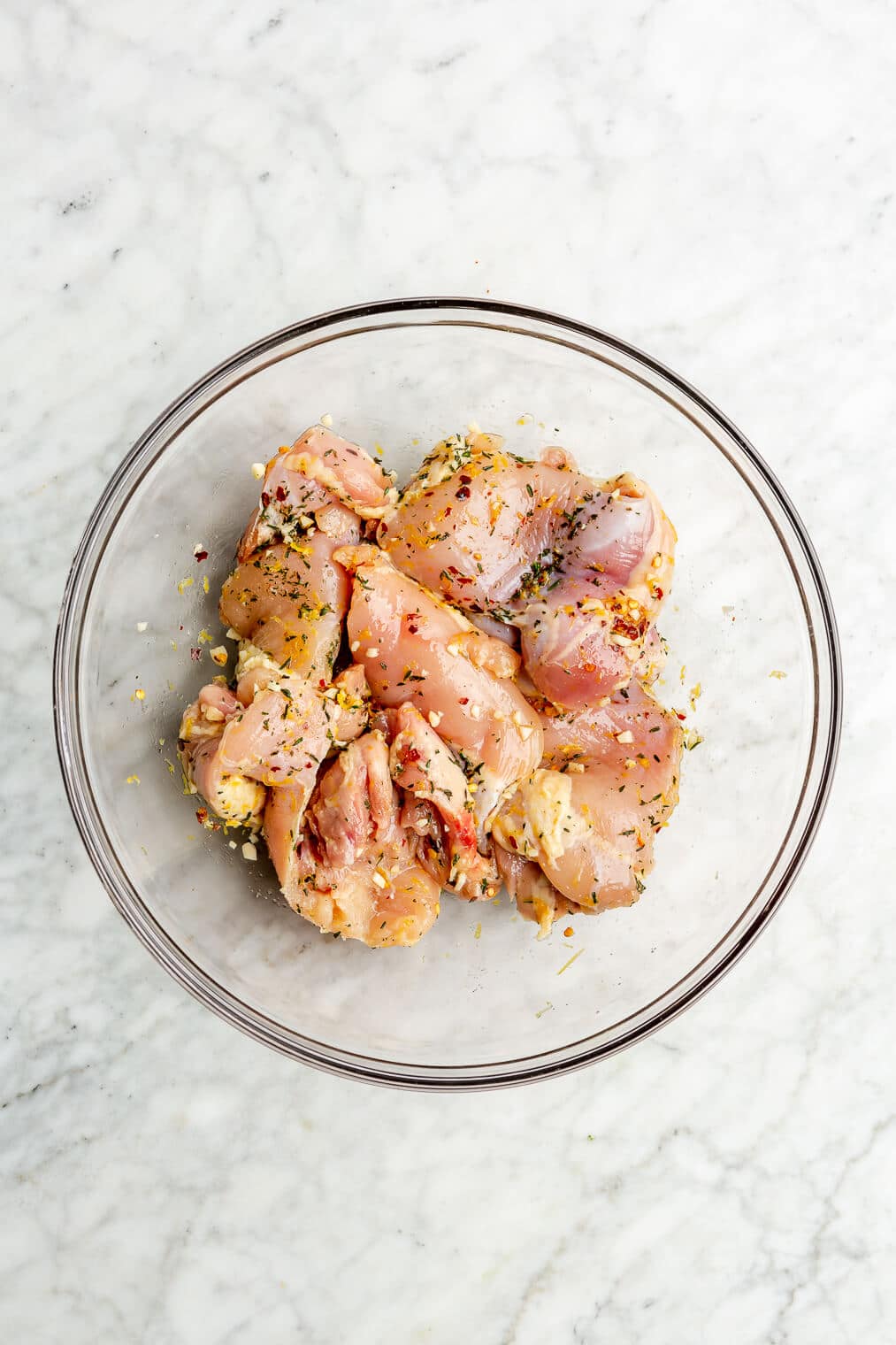 Top down of a glass bowl with chicken thighs tossed with lemon zest, herbs, and garlic on a grey and white marble surface.