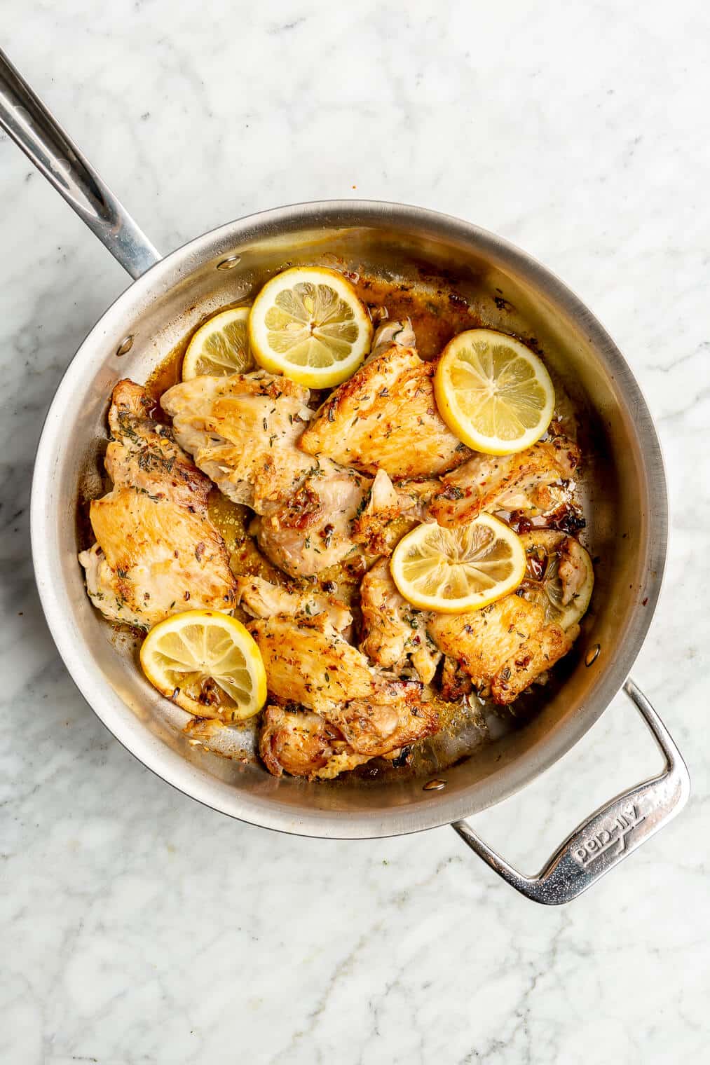 Top down of a skillet with browned chicken thighs and slices of lemons on a grey and white marble surface.