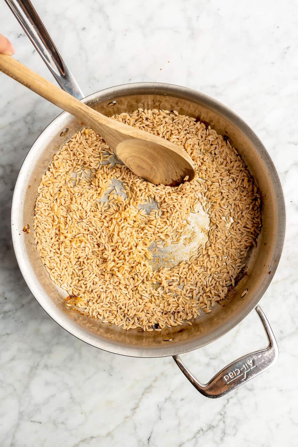 Top down photo of an all-clad skillet with orzo being toasted and stirred in butter with a wooden spoon on a grey and white marble surface.