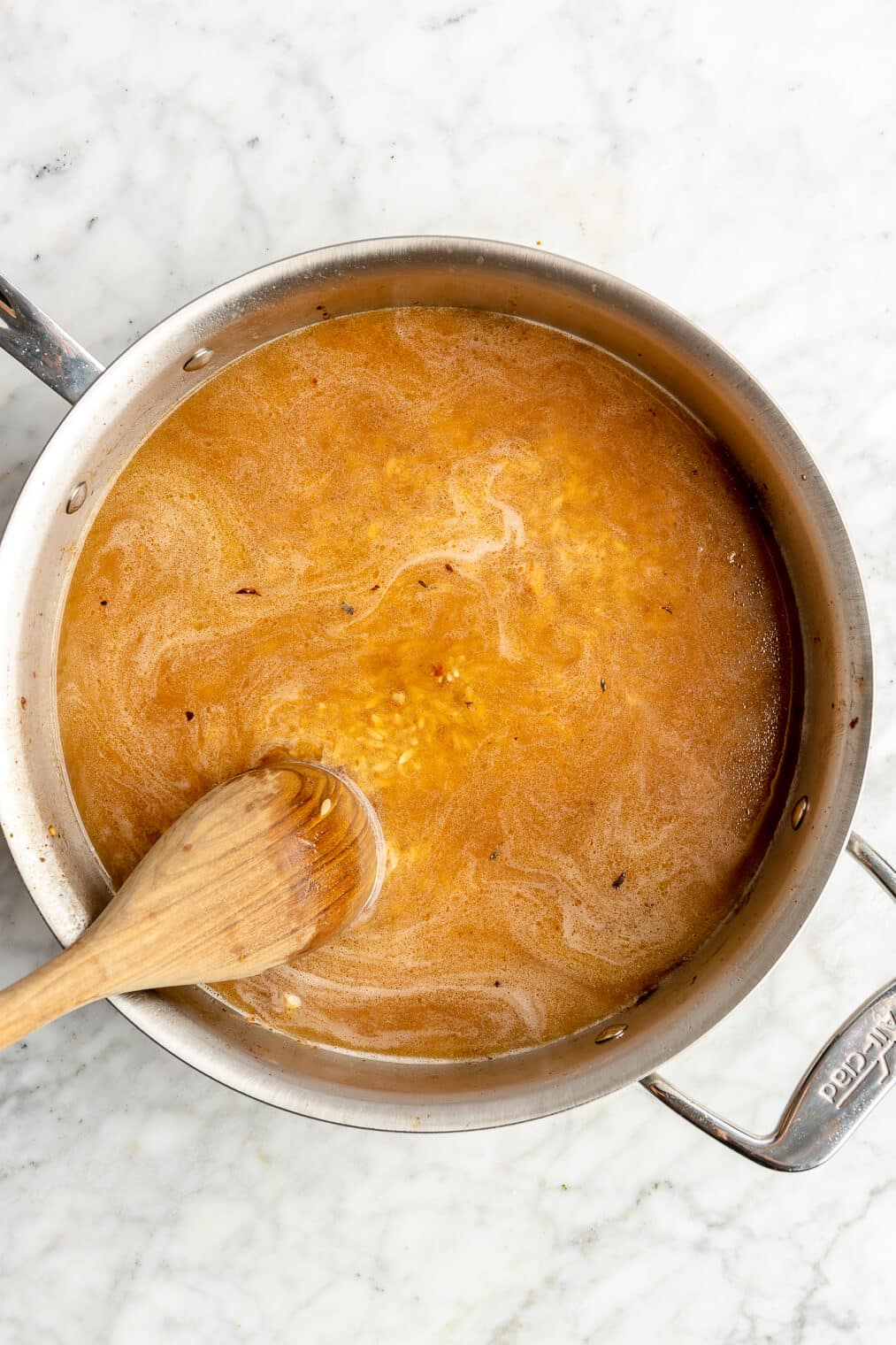 A wooden spoon stirring broth in an all-clad skillet with orzo on a grey and white marble surface.