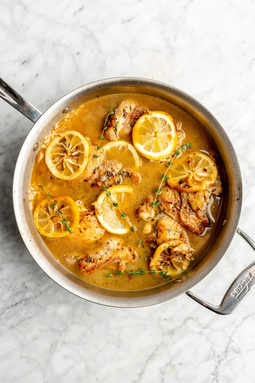 Top down view of browned chicken thighs and sliced lemons nestled in a skillet with broth on a grey and white marble surface. There are sprigs of thyme gentle placed over top of the chicken.