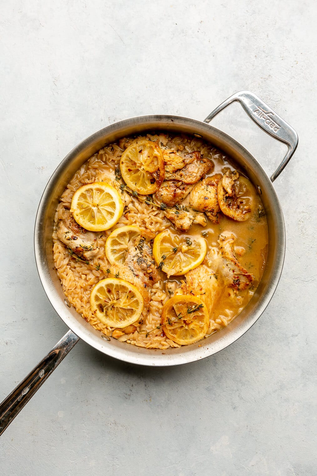 Cooked orzo, chicken thighs, lemon slices, and thyme with a little broth in an all-clad skillet on a grey surface.