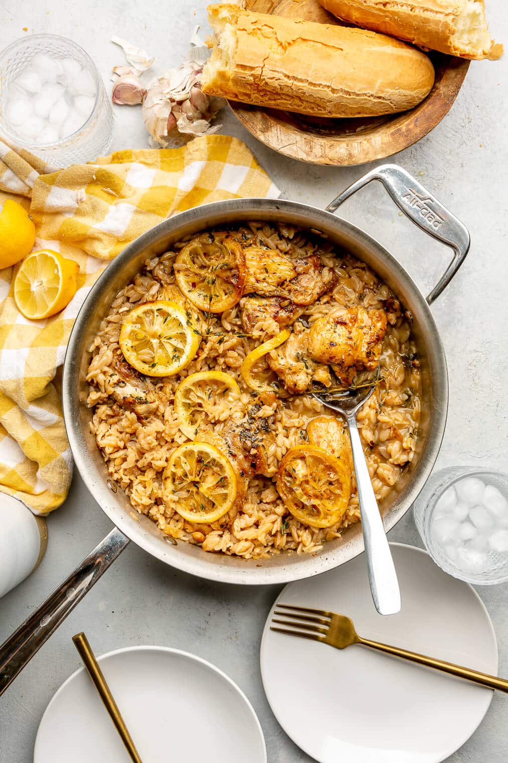 An all-clad skillet with orzo, chicken thighs, lemon slices, and thyme with a silver serving spoon on a grey surface. There are two white plates with gold forks, a couple glasses with ice water, a wooden bowl with a French loaf of bread ripped in half, a bulb of garlic, and a yellow and white checkered linen on the surface.