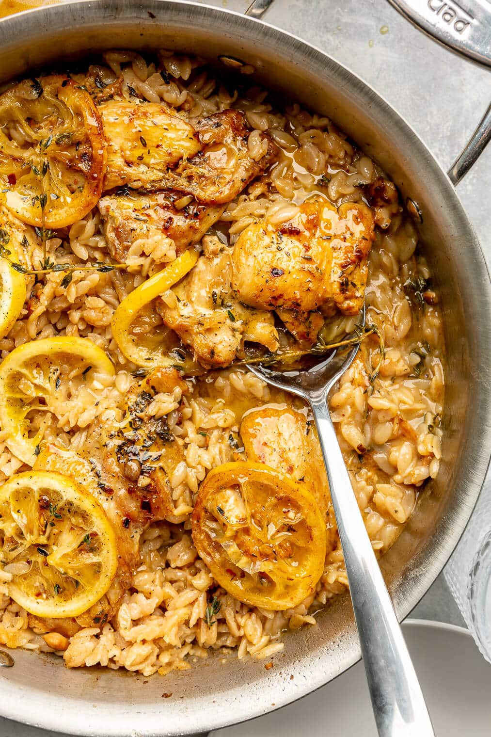 An all-clad skillet with orzo, chicken thighs, lemon slices, and thyme with a silver serving spoon on a grey surface.