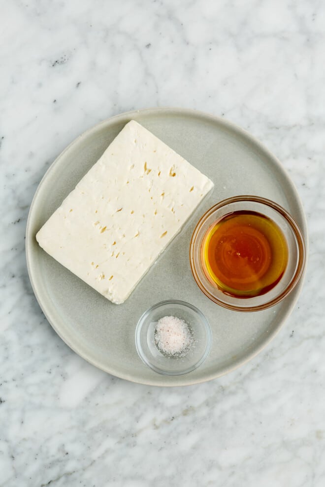 Whipped honey feta ingredients on a round, grey plate on a grey and white marble surface.