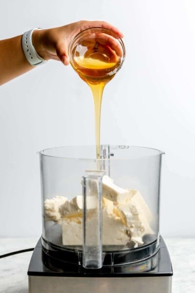 A hand pouring honey over top of blocks of feta in a food processor.