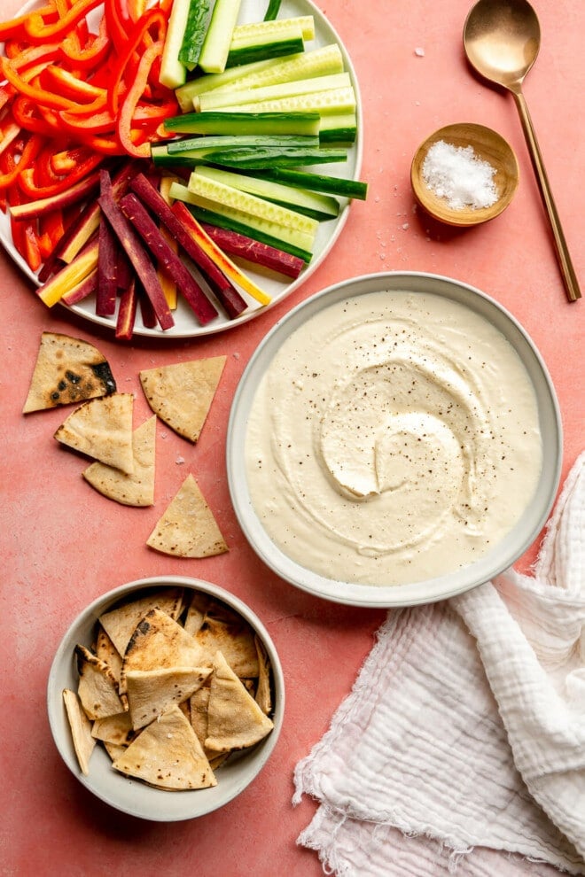 A white bowl of honey whipped feta topped with cracked black pepper. There is a small grey bowl of toasted pita bread, a plate with sliced red bell pepper, sliced cucumber, and sliced purple carrots and a small wooden bowl with flaky sea salt next to a copper spoon. All are sitting on a pink surface.