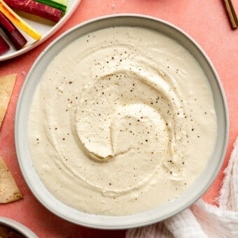 A white bowl with whipped honey feta topped with cracked black pepper on a pink surface. There is a plate with sliced vegetables to the top left corner and a small, wooden bowl with flaky sea salt to the top right. There is a white linen draped to the bottom and a small bowl of toasted pita bread.