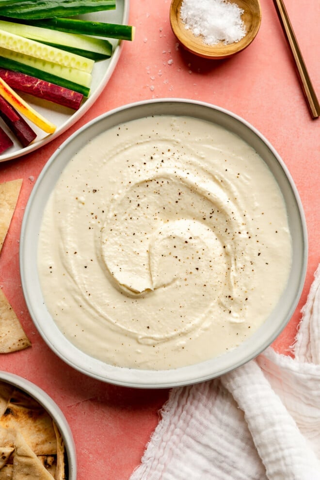 A white bowl with whipped honey feta topped with cracked black pepper on a pink surface. There is a plate with sliced vegetables to the top left corner and a small, wooden bowl with flaky sea salt to the top right. There is a white linen draped to the bottom and a small bowl of toasted pita bread.