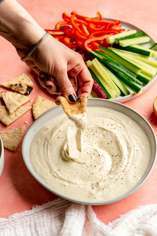 A hand dipping a toasted pita into the bowl of honey whipped feta. There is a plate of sliced vegetables in the background and a white linen draped to the front.