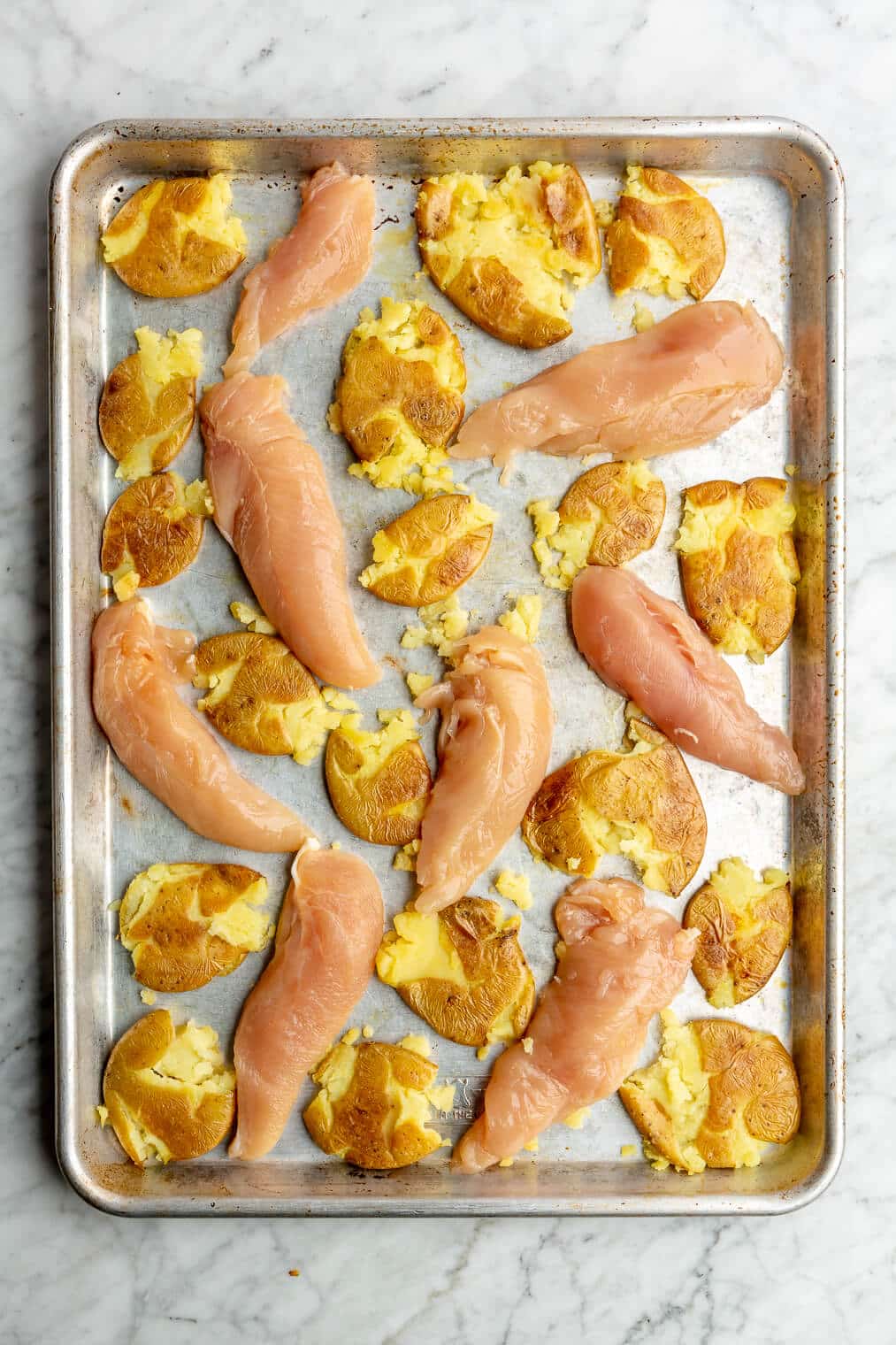 Smashed potatoes and raw chicken tenders on a sheet pan.
