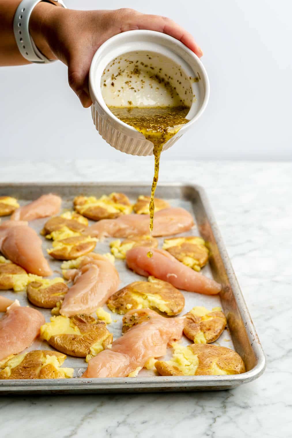 A person pouring Greek dressing over a sheet pan of raw chicken tenders and smashed potatoes.