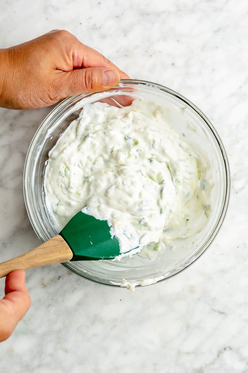 A person using a rubber spatula to stir together a homemade tzatziki sauce in a small glass bowl.