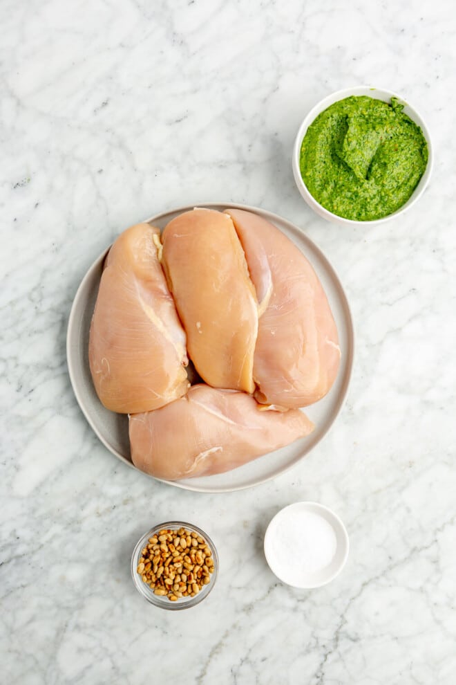 All of the ingredients for pesto chicken salad (chicken breasts, pine nuts, pesto, and salt) on a marble surface.