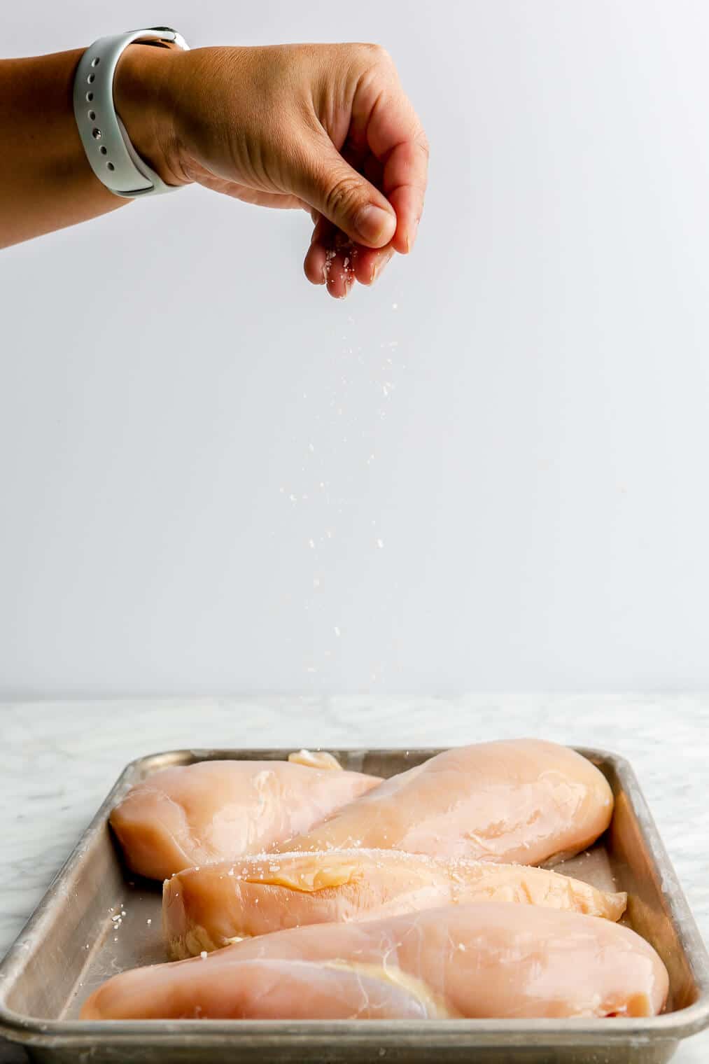 A person sprinkling sea salt over raw chicken breasts on a sheet pan.