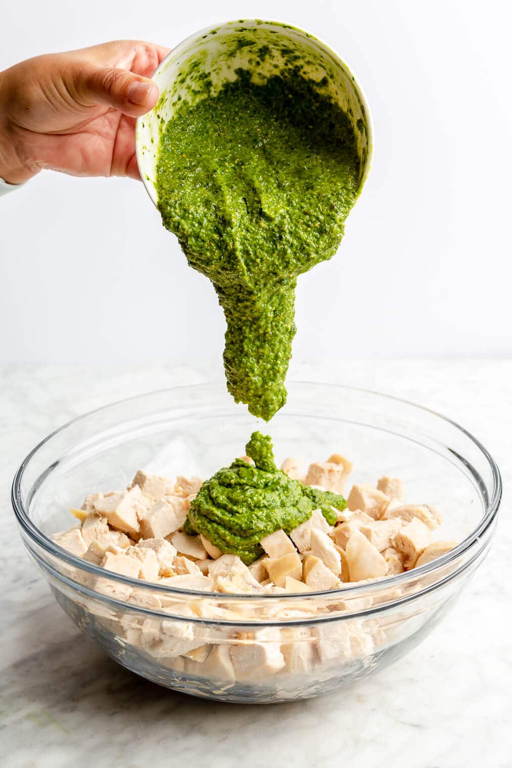 A person pouring a bowl of homemade dairy-free pesto into a bowl of cubed chicken breasts.