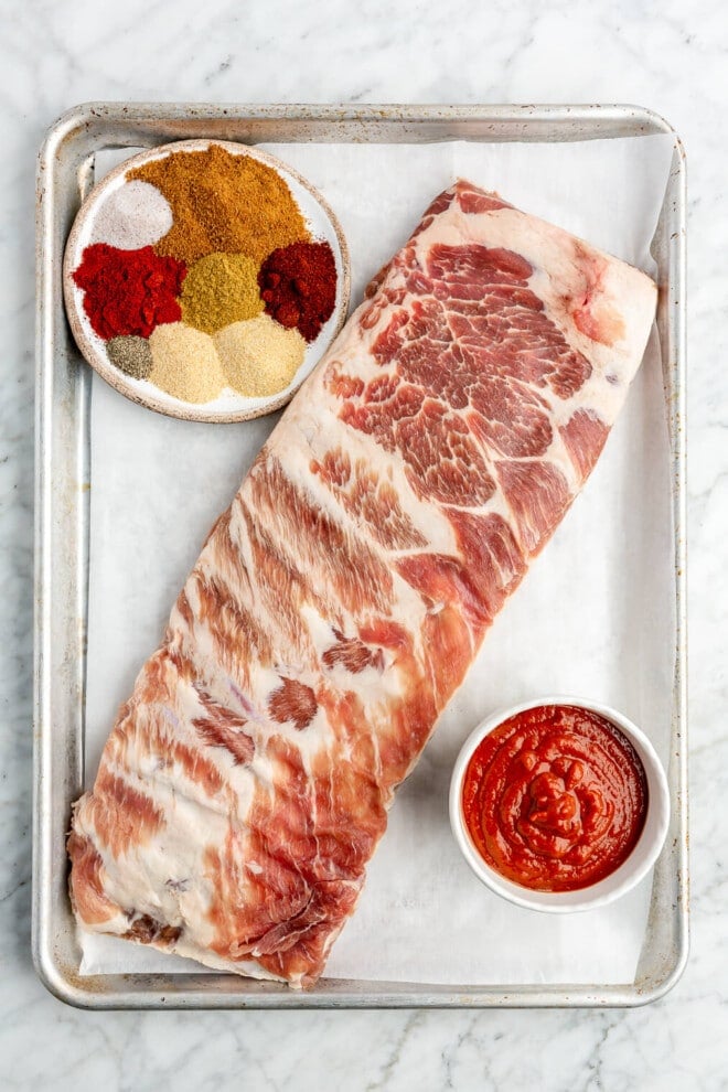 All of the ingredients for pork spare ribs on a sheet pan that's sitting on a marble surface.
