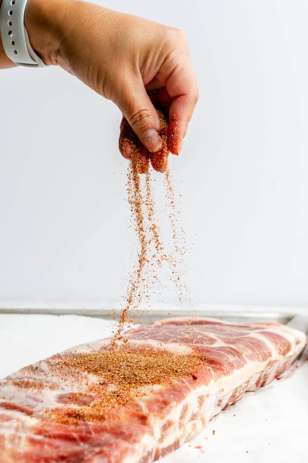 A person sprinkling BBQ spice rub onto pork spare ribs.