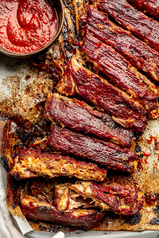 An up close shot of BBQ pork spare ribs on a sheet pan next to a small bowl of BBQ sauce.