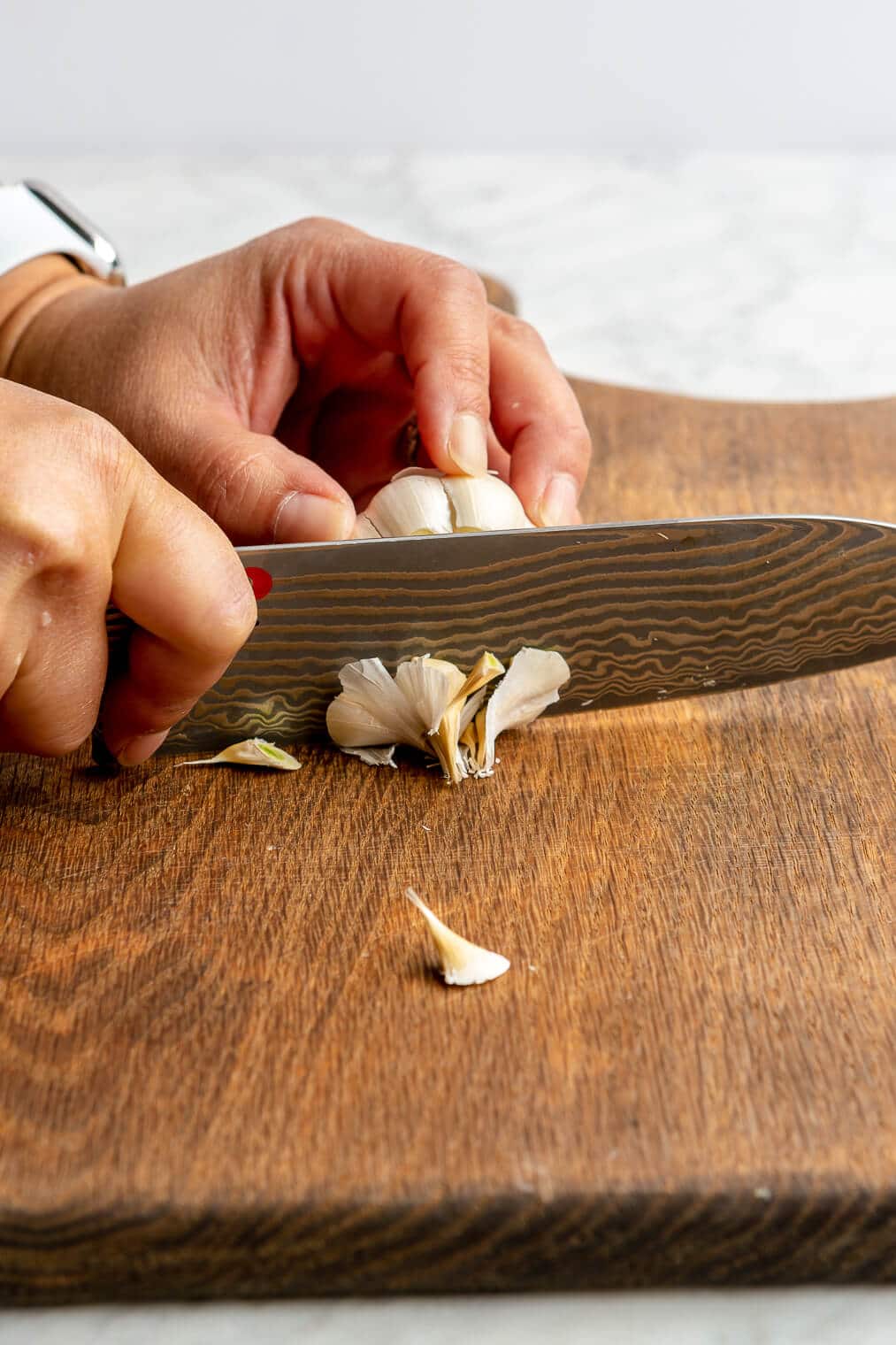 A person chopping off the tip of a bulb of garlic on a wooden cutting board.