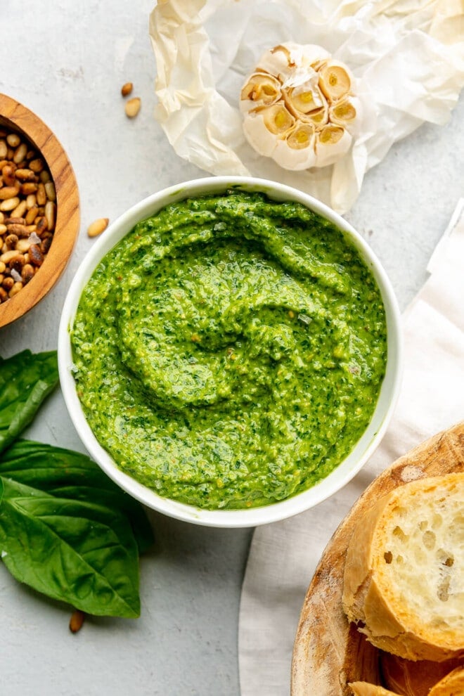 A bowl of dairy-free pesto on a light gray surface next to a bowl of toasted pine nuts, roasted garlic, and toast.
