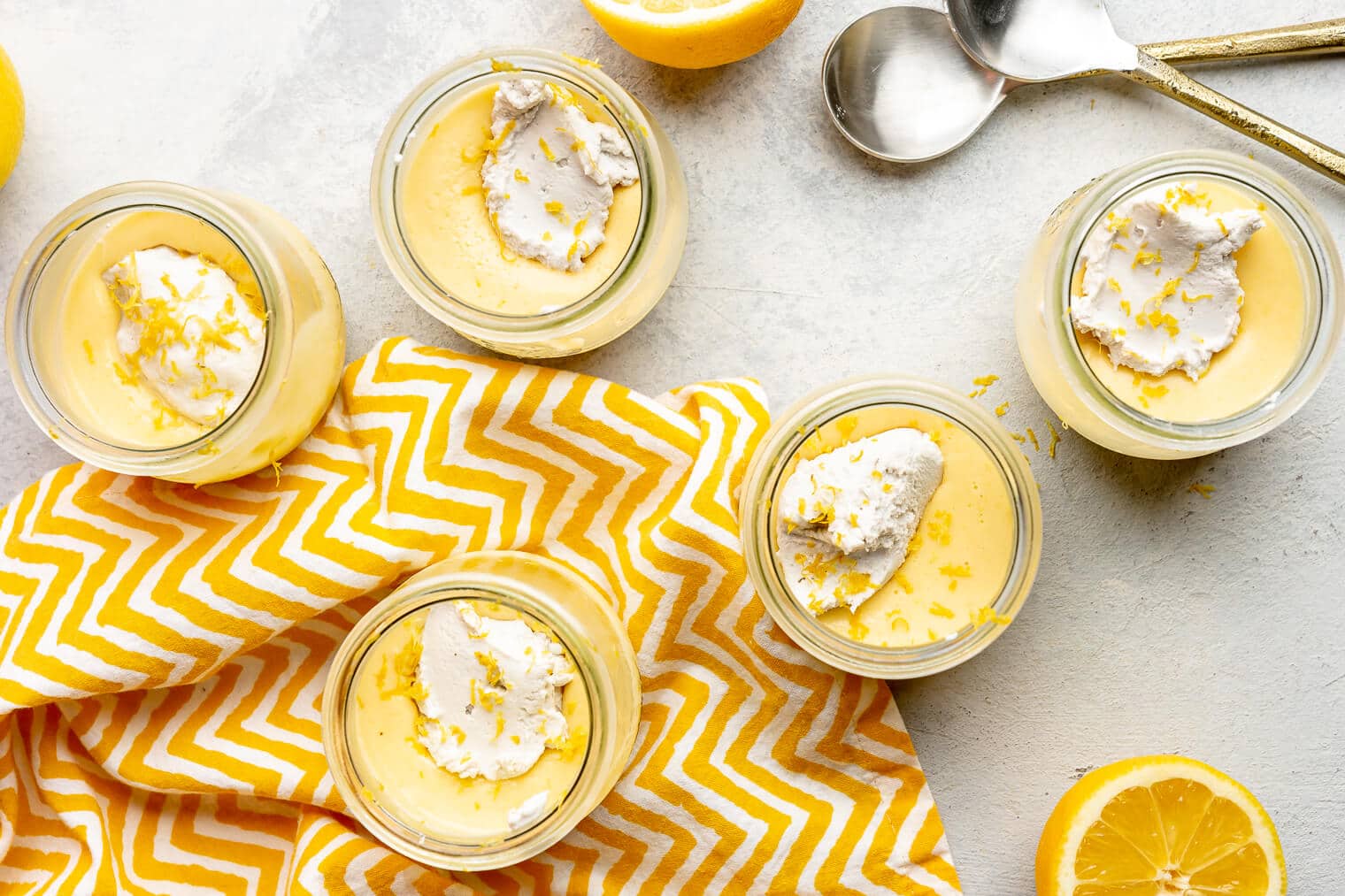 A top view of 5 jars of lemon curd, each topped with coconut whipped cream and lemon zest.