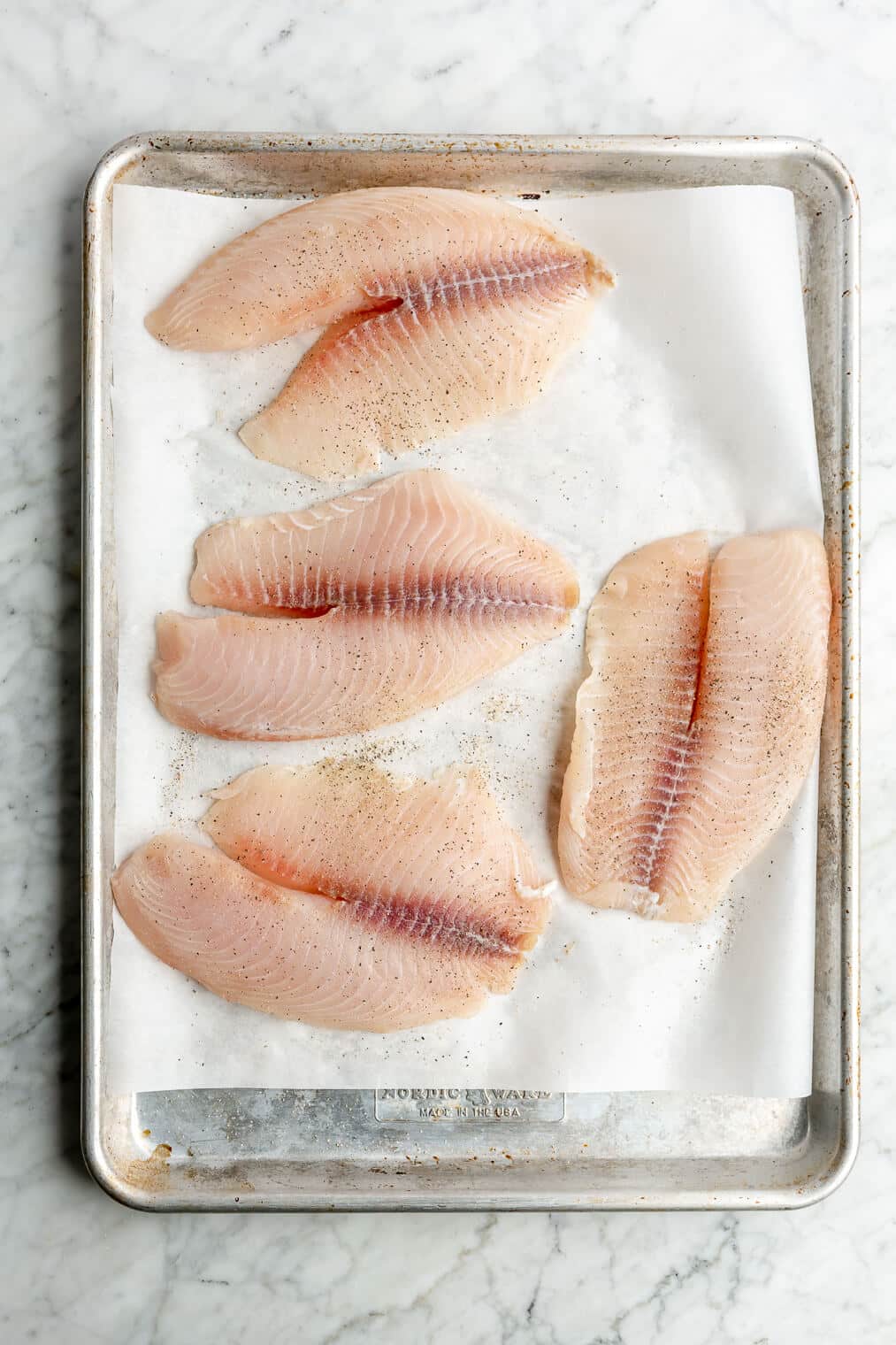 Top view of 4 raw tilapia filets on a parchment paper lined sheet pan.