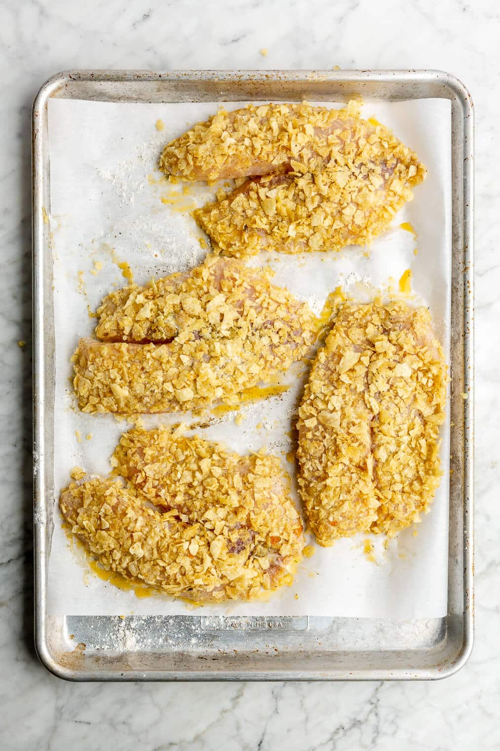 A parchment paper lined sheet pan with 4 raw potato chip crusted tilapia filets on it.