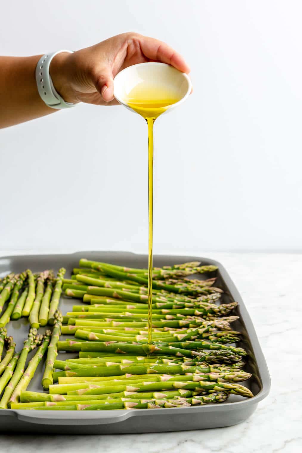 A person pouring a small bowl of olive oil over asparagus on a sheet pan.
