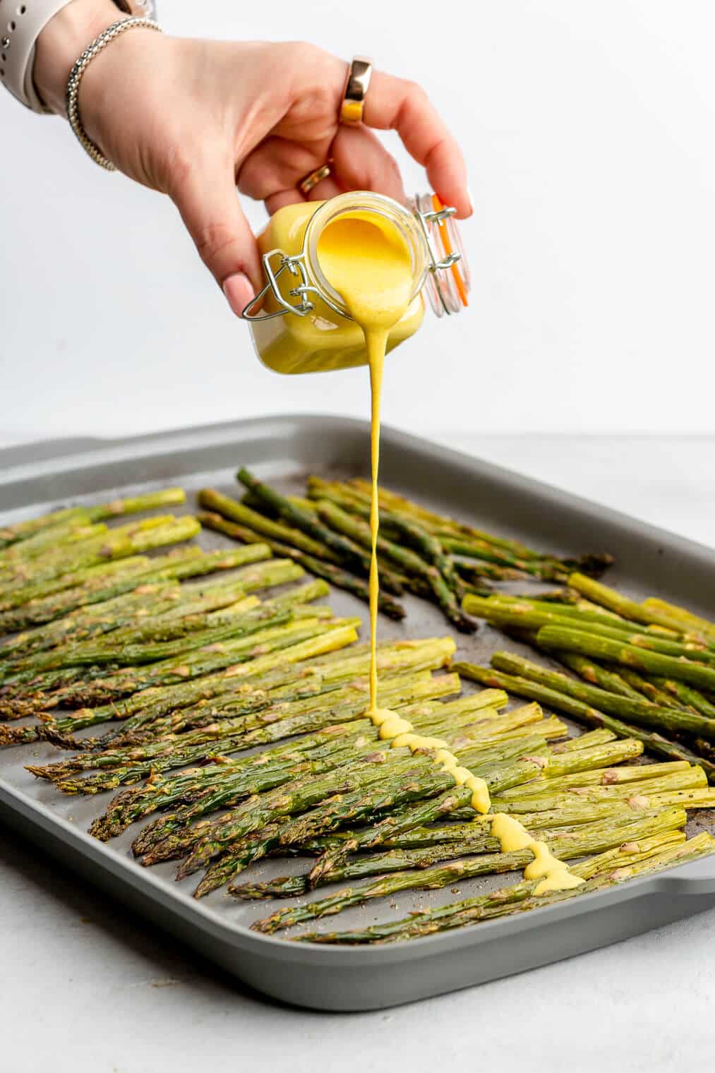 A person pouring bright yellow lemon cardamom sauce over a sheet pan of roasted asparagus.