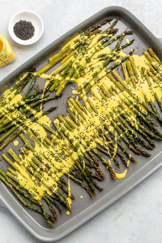 Roasted asparagus on a sheet pan topped with a bright yellow lemon cardamom sauce and black sesame seeds.