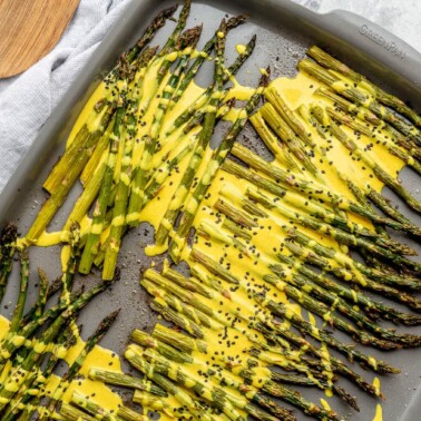 Roasted asparagus on a sheet pan topped with a bright yellow lemon cardamom sauce and black sesame seeds.