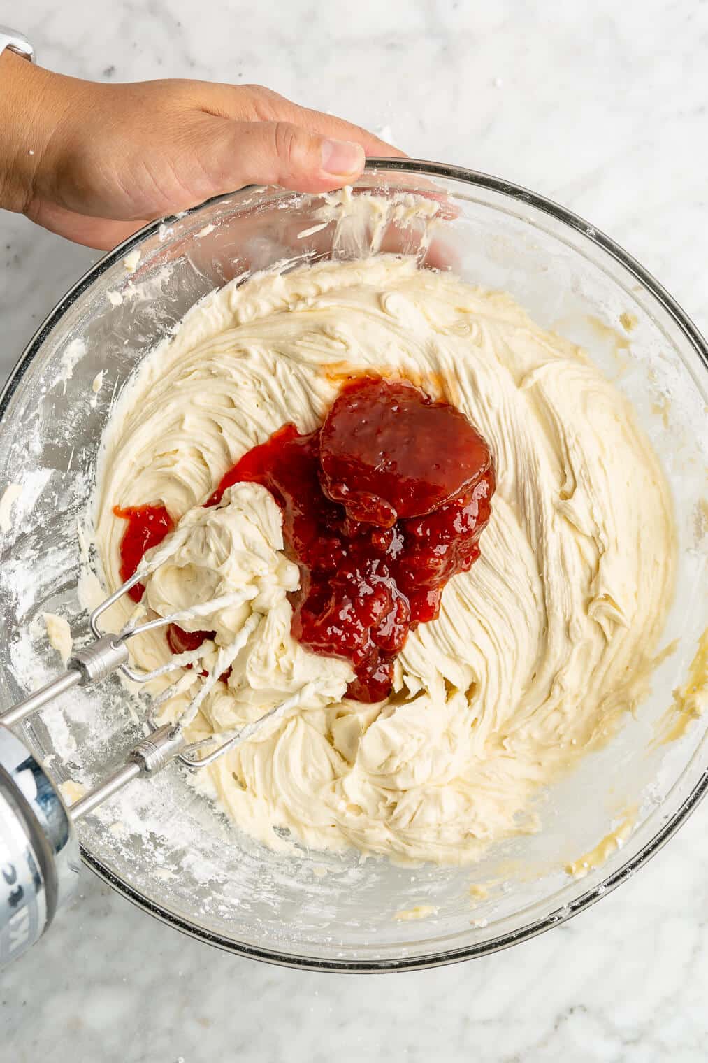 A large bowl of cream cheese frosting with strawberry jam being mixed in with an electric hand mixer.