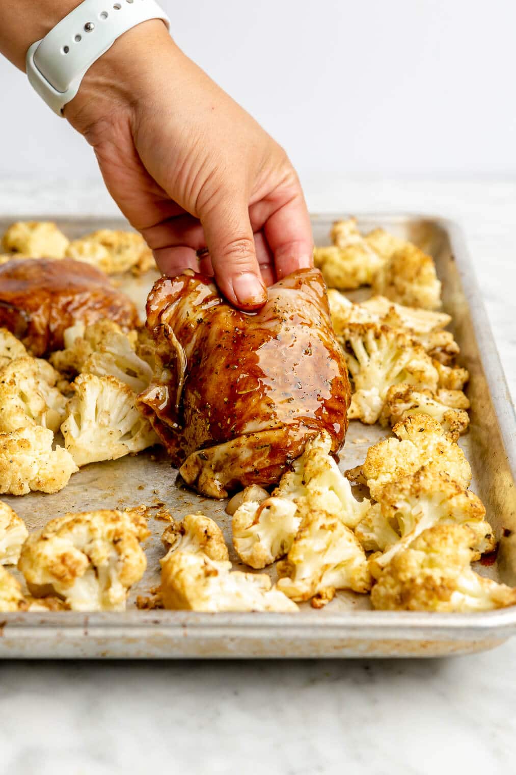 A person placing a raw balsamic coated chicken thigh onto a sheet pan with cauliflower.