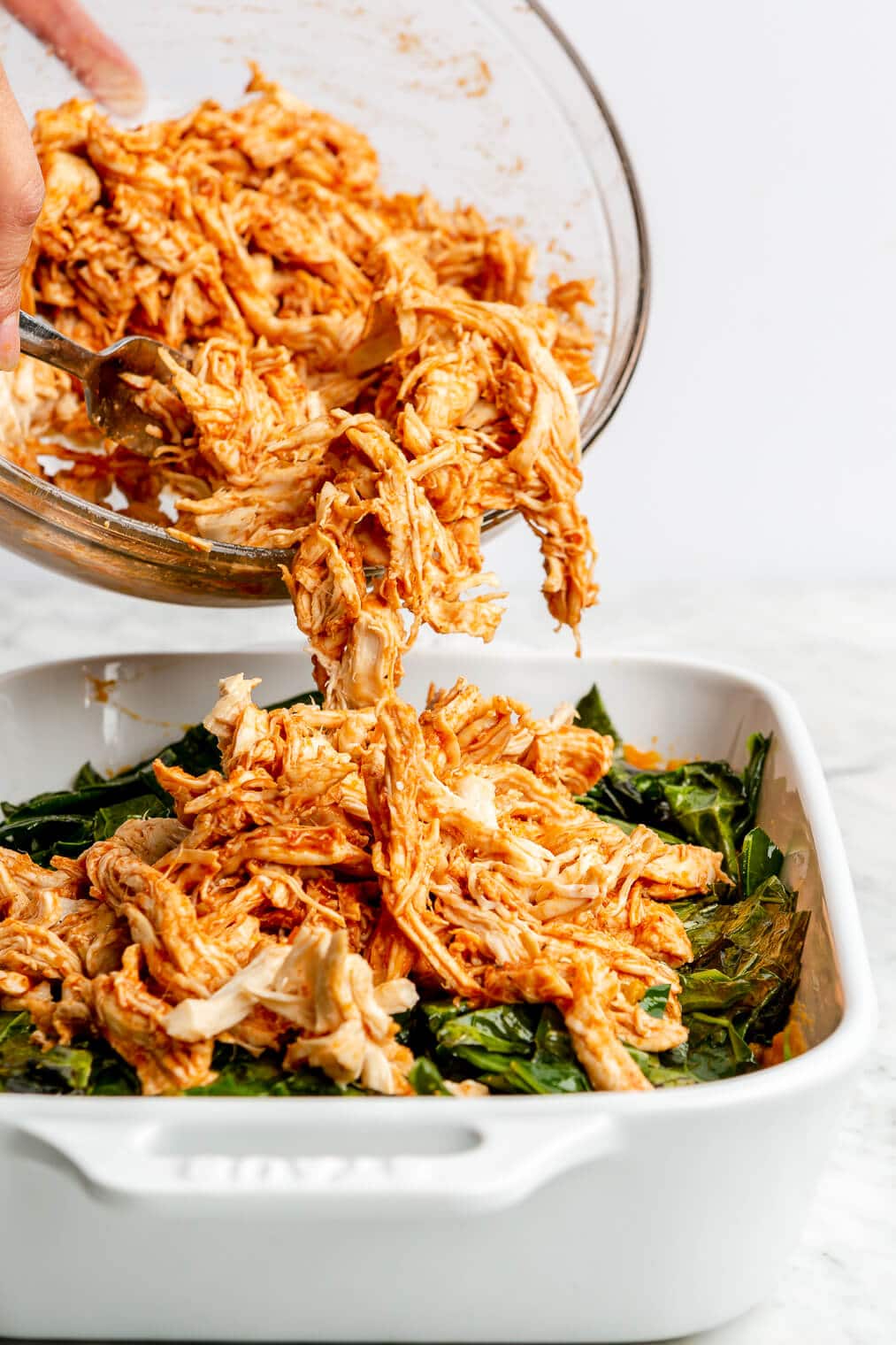 A person spooning shredded BBQ chicken over sweet potato and kale in a casserole dish.