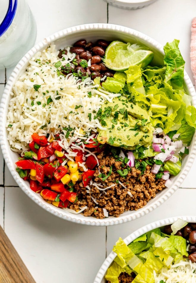 An assembled burrito bowl with rice, beans, peppers, onions, corn, ground beef, lettuce, guacamole, and cheese.