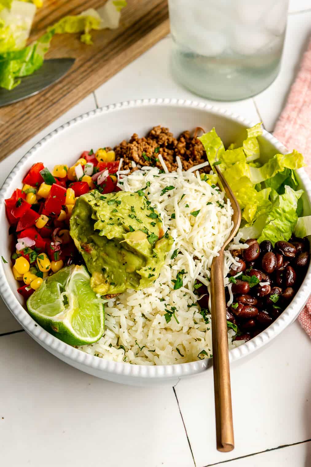 An assembled burrito bowl with rice, beans, peppers, onions, corn, ground beef, lettuce, guacamole, and cheese.