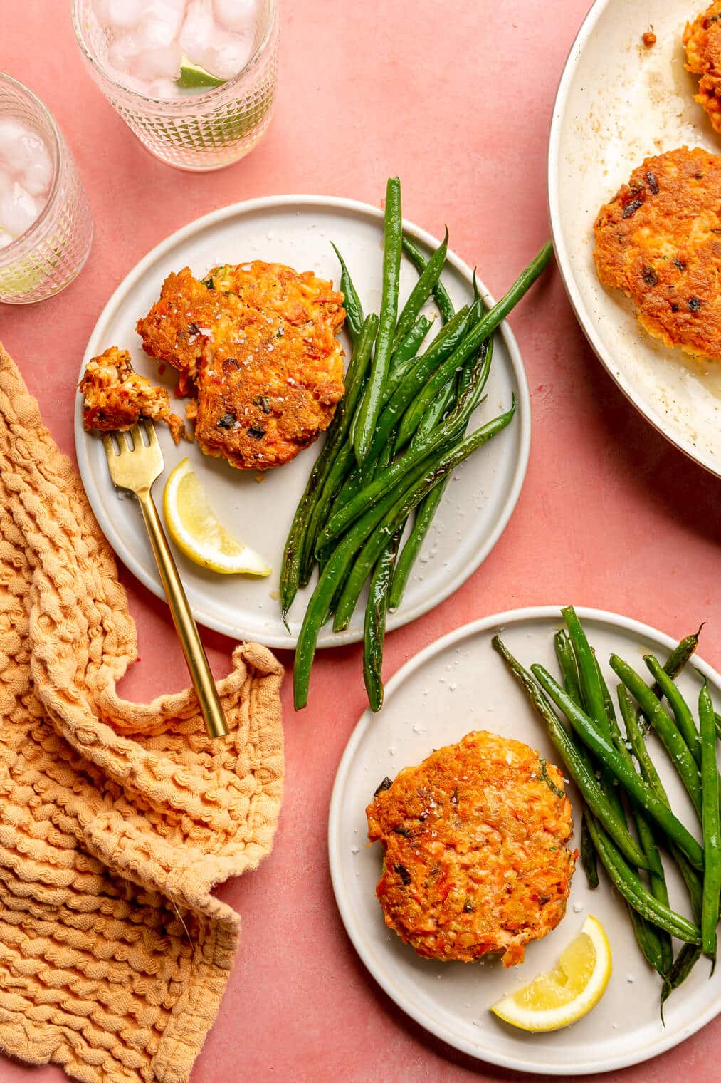 Two servings of salmon cakes and sauteed green beans on plates next to a skillet with more salmon cakes and two glasses of water.