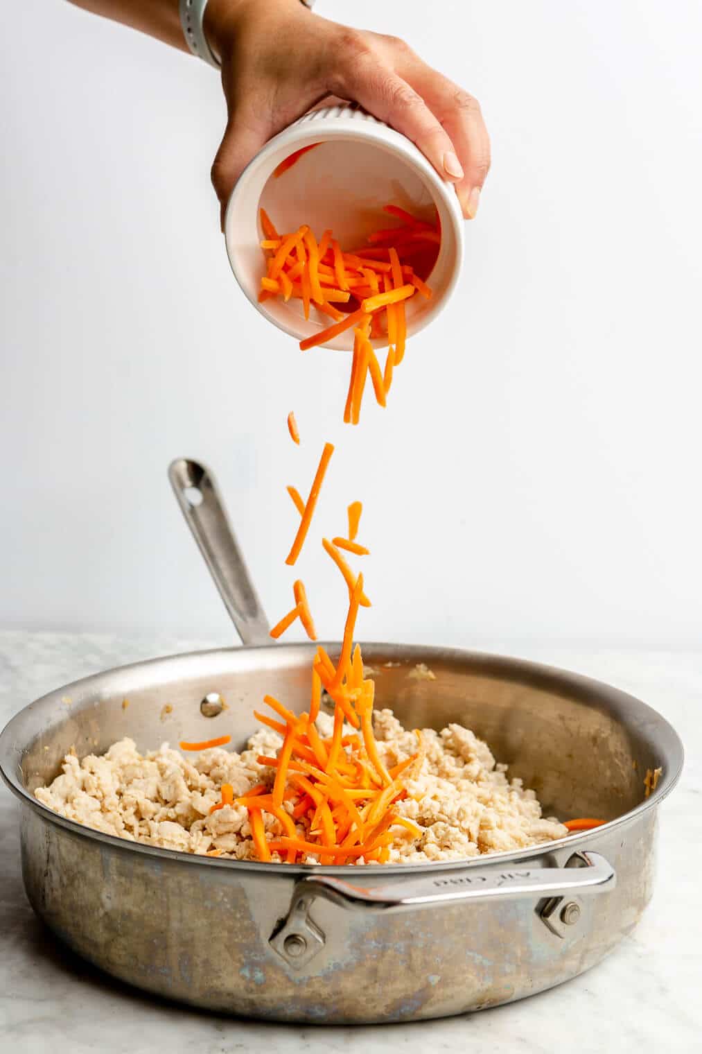 A person adding shredded carrots into a large skillet with cooked and crumbled ground turkey.